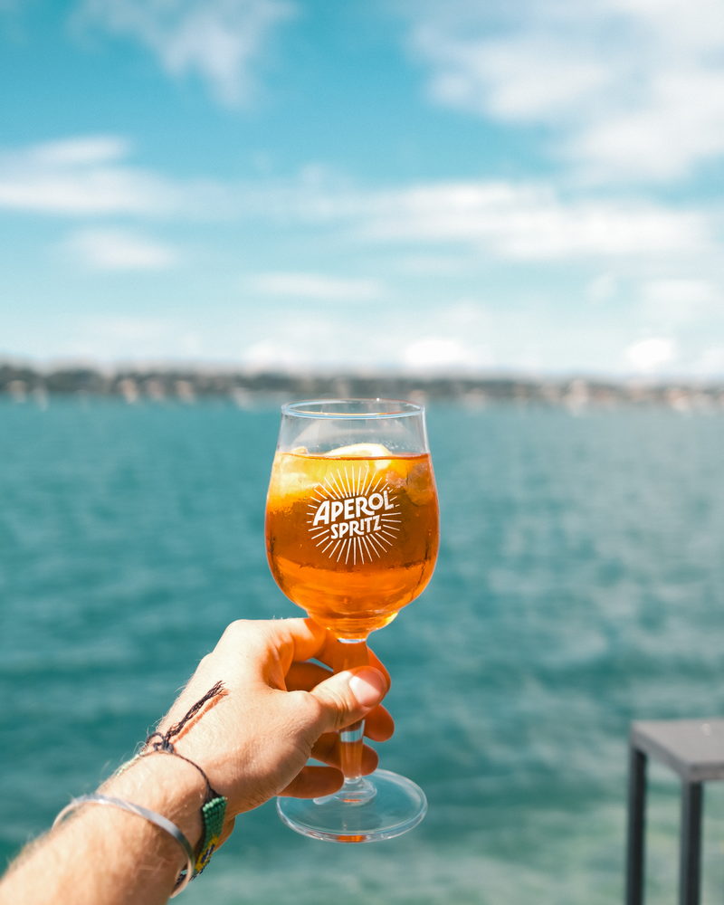 Ein Glas Aperol Spritz, in die Höhe gehalten vor dem türkisfarbenen See