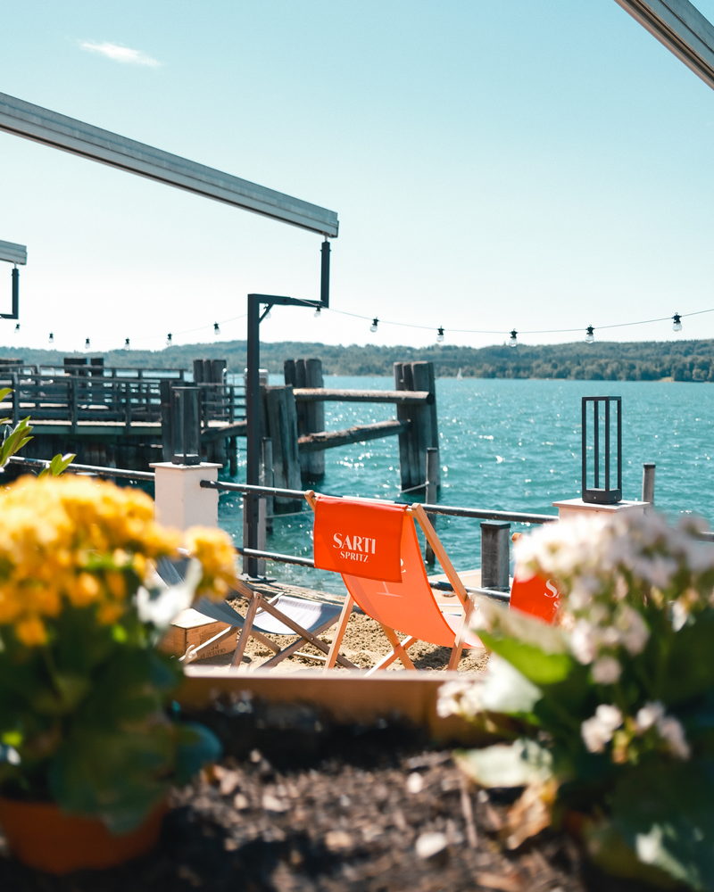 Orange Liegestühle im Sand am Ufer, dahinter Bootssteg und See