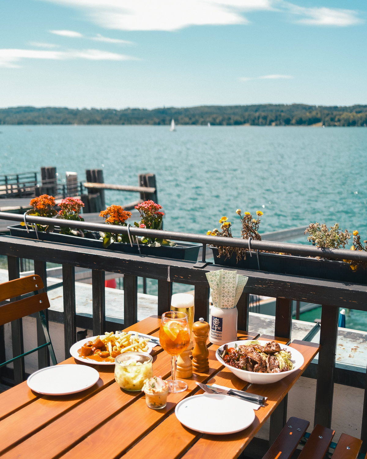 Gedeckter Tisch auf der Restaurantterrasse mit Currywurst, Salat, Spritz und Bier, dahinter der Starnberger See mit Segelboot